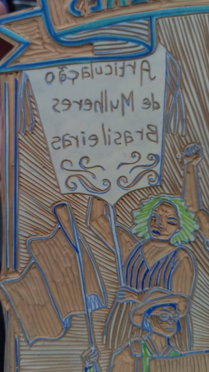 Fotografia com o detalhe da matriz de xilogravura do Manifesto 8 de março de 2022. Uma placa de linóleo de cor amarronzada, com vincos que ressaltam os desenhos. No detalhe, uma mulher segura um estandarte da Articulação de Mulheres Brasileiras e uma mulher com chapéu que segura uma bandeira.