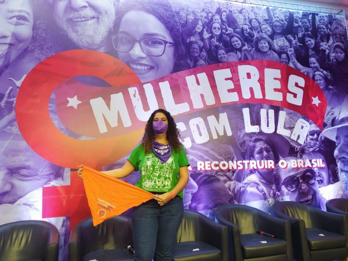 "Transformando o Mundo pelo Feminismo" e a logo da AMB, diante de um grande telão com imagem em azul de Lula com diversas mulheres e escrito: "Mulheres com Lula".