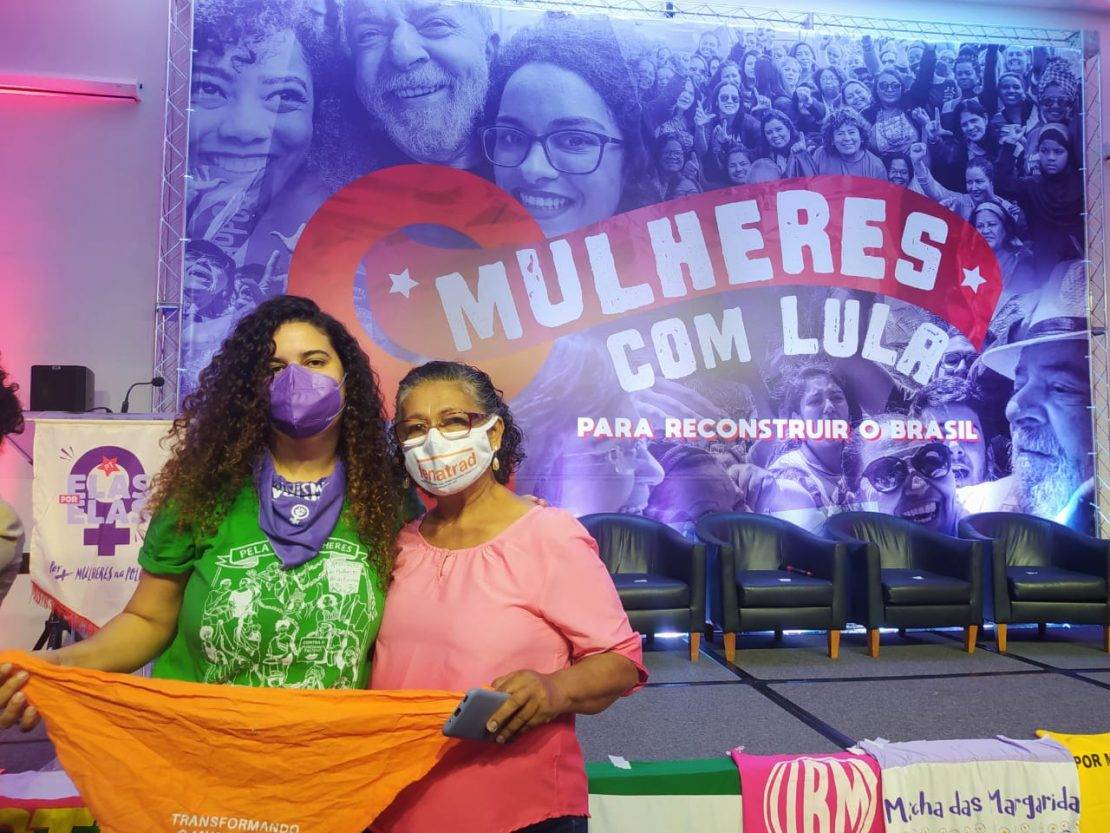 Fotografia de Elisa Aníbal (AMB) e Luiza Batista (FENATRAD) em evento com um grande telão ao fundo com imagem em tom de roxo de Lula com diversas mulheres e escrito: "Mulheres com Lula". As duas seguram, juntas, uma bandana laranja escrita: "Transformando o Mundo pelo Feminismo" e a logo da AMB