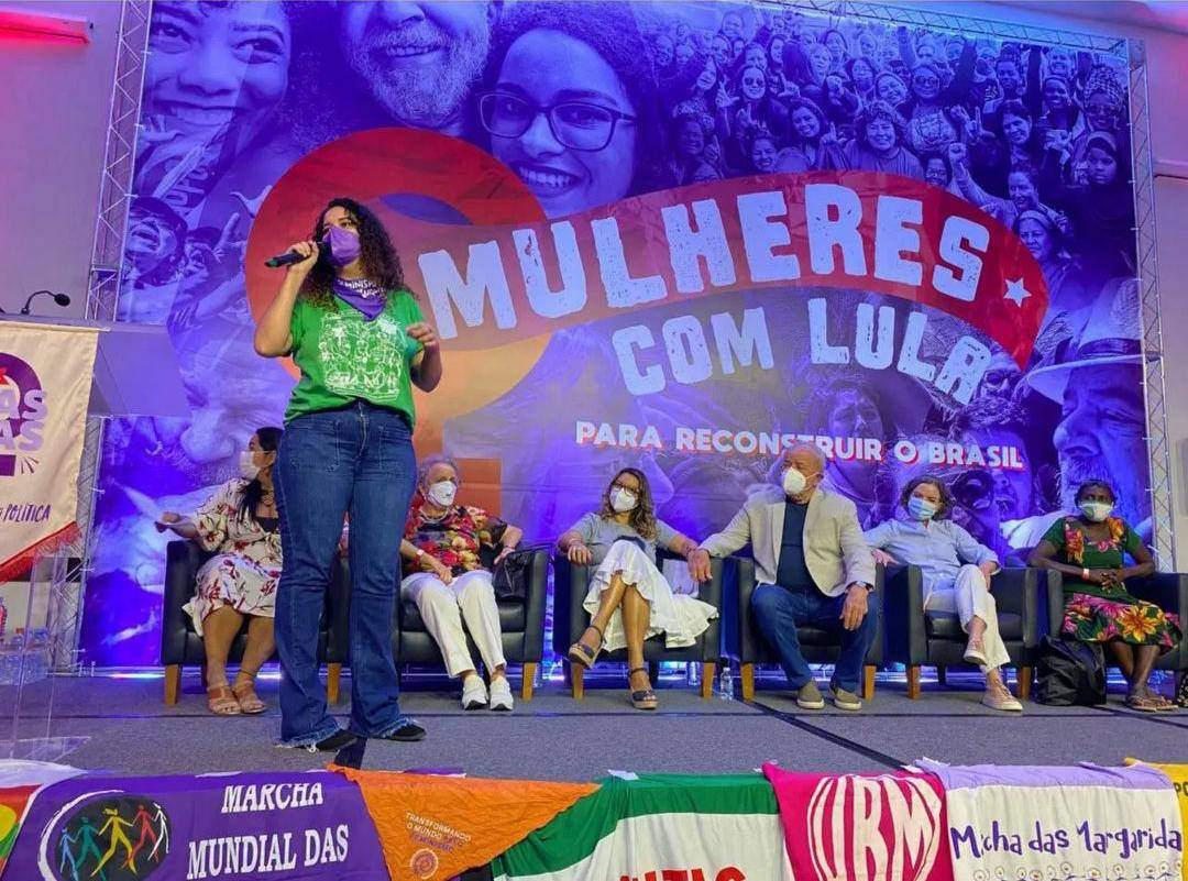 Fotografia da representante da AMB, Elisa Aníbal, falando com o microfone nas mãos, diante de um grande telão com imagem em azul de Lula com diversas mulheres e escrito: "Mulheres com Lula".