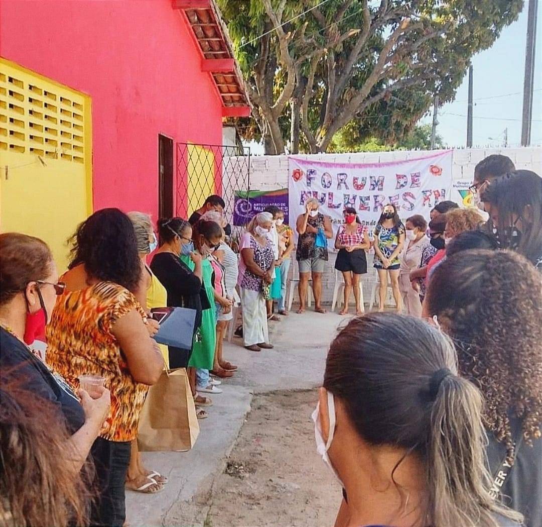 Diante de uma casa com fachada vermelha e janelas amarelas, na calçada, integrantes do Fórum de Mulheres do Rio Grande do Norte  estão reunidas em um semicírculo.