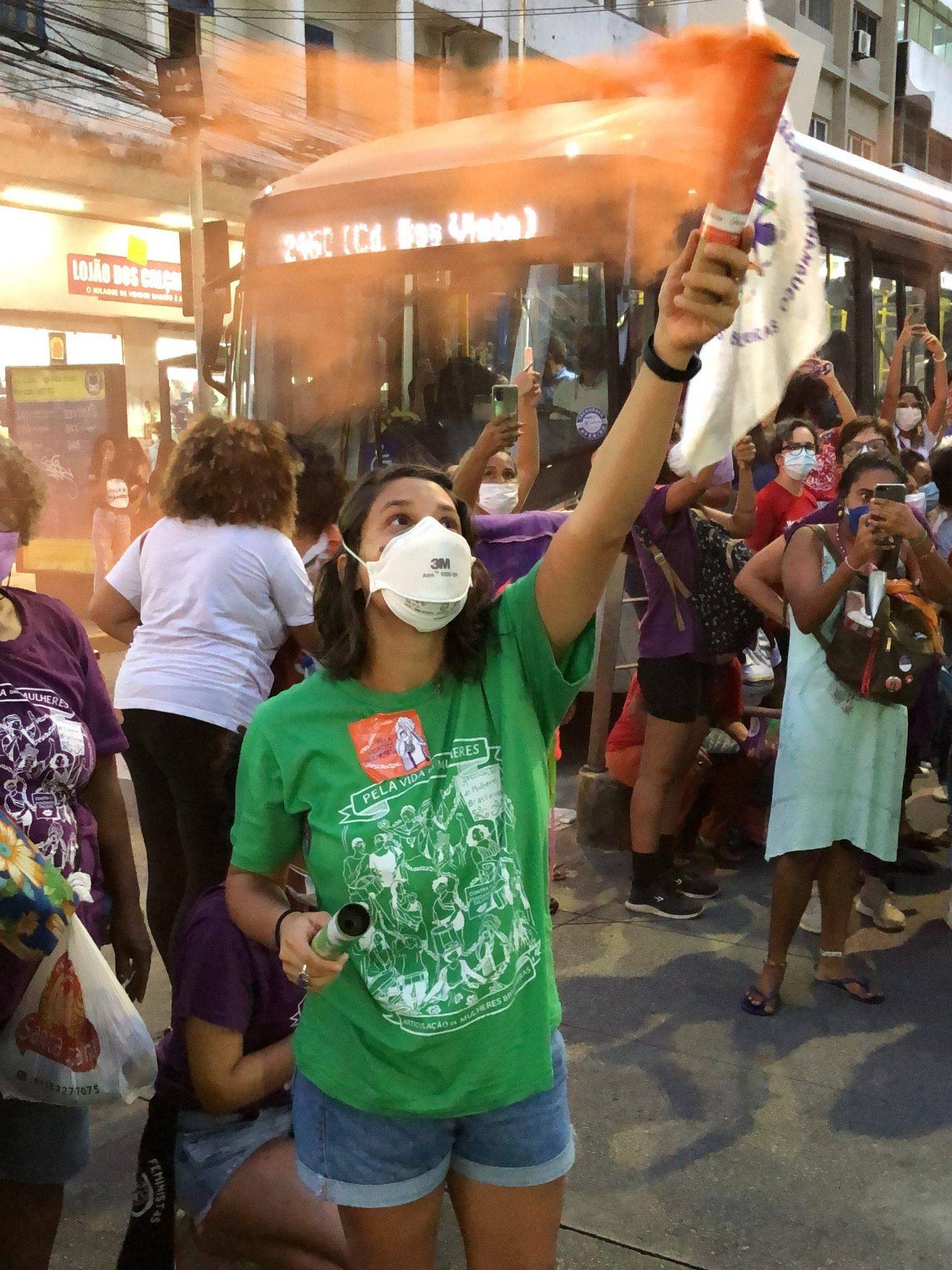 Em ato de rua mulher ergue sinalizador de fumaça laranja, ela usa máscara contra a COVID-19 e camiseta verde com as identidade visual da AMB.