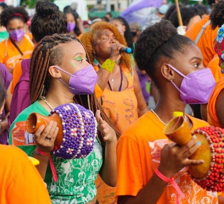 Em ato de rua duas meninas negras, em primeiro plano, tocam agbê/ xequere. Atrás delas uma mulher negra canta ao microfone. As cores laranja, roxo e verde são muito presentes, nas camisas, instrumentos e máscaras.