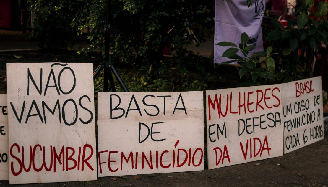 No chão há quatro placas de madeira clara estão escritas frases em tinte preta e vermelha: "Não vamos sucumbir", "Basta de feminicídio", Mulheres em defesa da vida" e "Brasil um caso de feminicídio a cada 6 horas."
