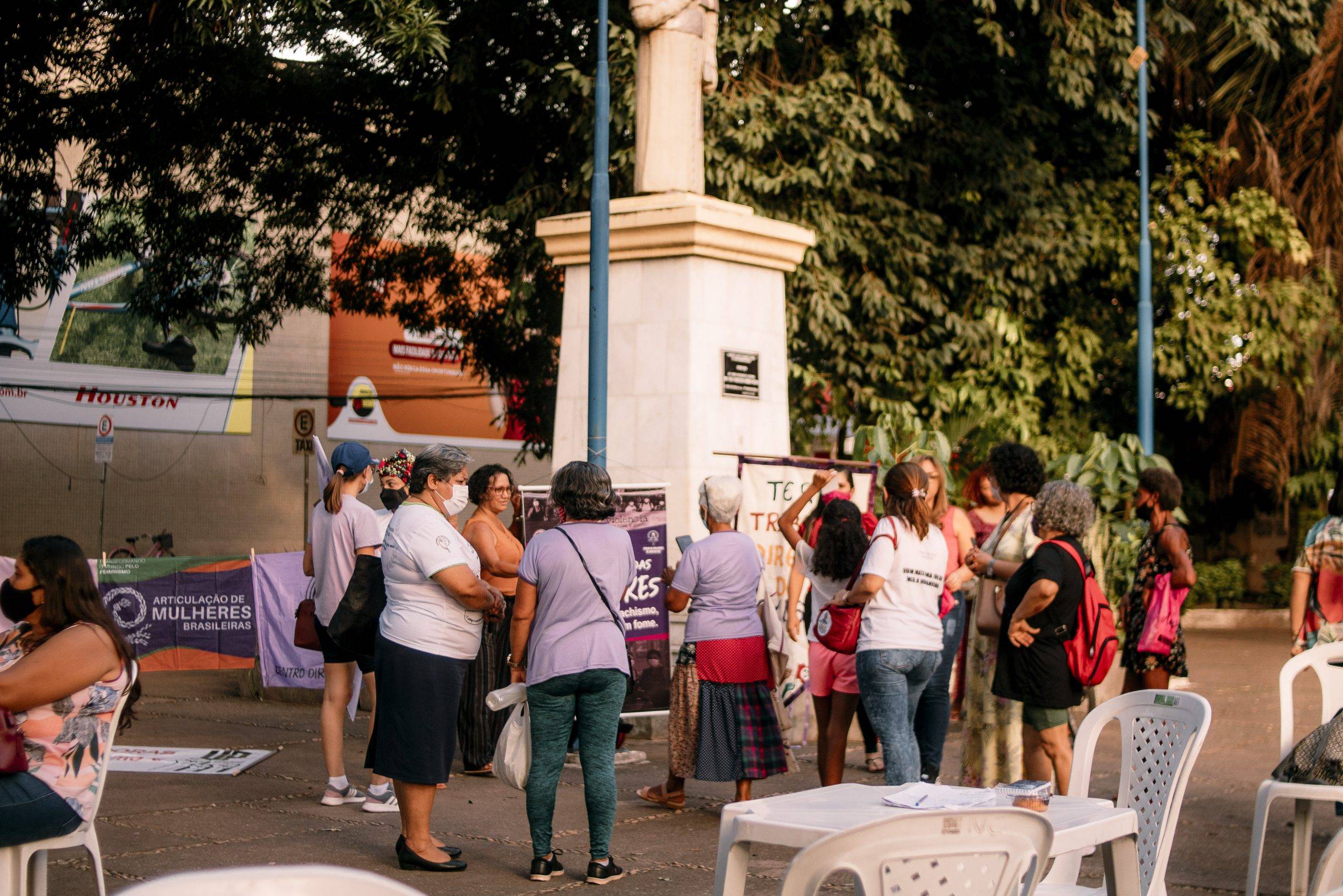 Em volta de estátua, na Praça de Fátima, há um varal onde estão penduradas várias bandeiras e faixas de movimentos. Várias mulheres diversas conversam por perto.