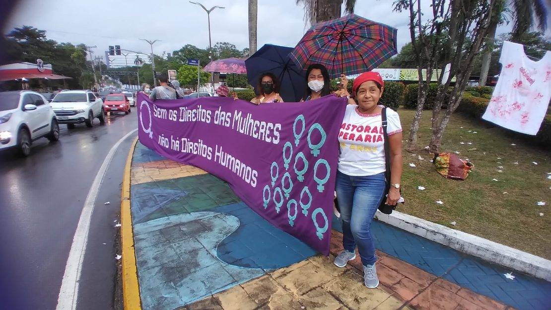 Em calçada de avenida agitada, algumas mulheres diversas seguram uma grande faixa roxa, com vários espelhos de vênus azuis, em letras brancas escrito: "Sem os direitos das mulheres não há direitos humanos."